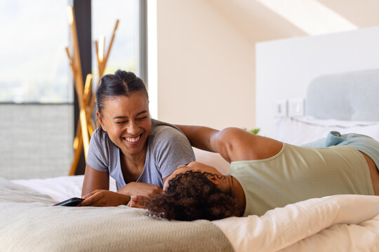 Happy biracial lesbian couple embracing and using smartphone on bed in bedroom - Powered by Adobe
