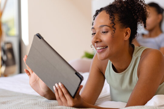 Happy biracial lesbian couple using tablet on bed in bedroom