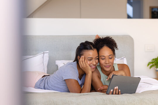 Happy biracial lesbian couple using tablet on bed in bedroom