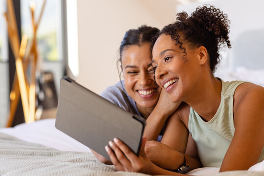 Happy biracial lesbian couple using tablet on bed in bedroom - Powered by Adobe