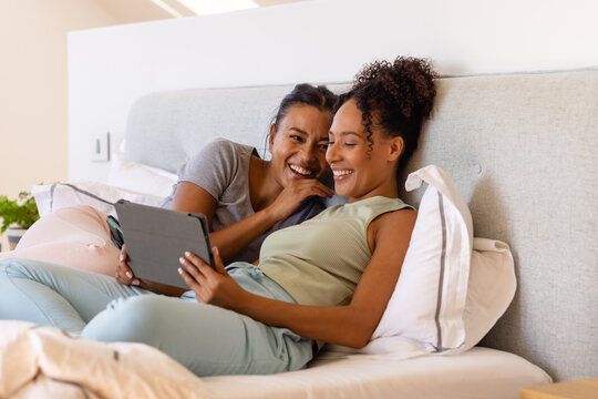 Happy biracial lesbian couple using tablet on bed in bedroom