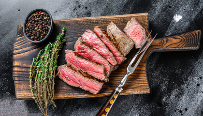 Sliced grilled roast beef with fork for meat on wooden cutting board. Black background. Top view.