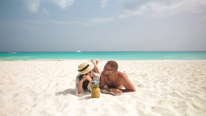 Latin couple lies and smiling on the white sand on the tropical beach. Pineapple cocktail and paradise pleasure against the background of the turquoise sea. - obrazy, fototapety, plakaty