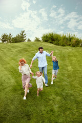 Happy couple, man and woman going for a walk in park with their little children, boy and girl, running, playing on grass. Concept of family, childhood and parenthood, fun, weekends, love, ad