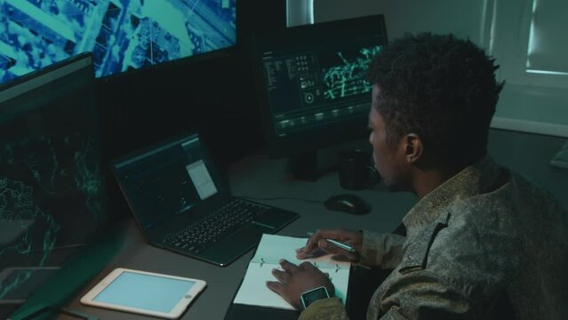 High angle medium shot of male African American military surveillance officer sitting at desk looking at screen with map tracking object, making notes and typing on keyboard