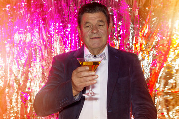 An elderly man with a glass of martini makes a celebratory toast against a shiny red background.