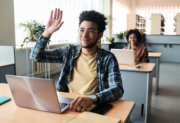 Student sitting at desk with laptop and raising his arm up to answer on question in classroom