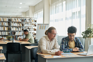 Mature teacher teaching language to foreign student while they sitting at desk in library