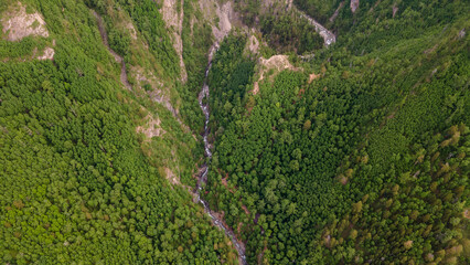 Aerial view of the high waterfall and fast mountain river in valley of the rocky mountain ranges, on which snow lies in places at summer cloud morning, yellow color