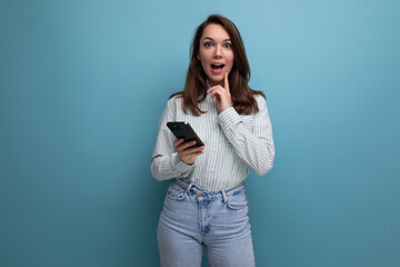 surprised european young brunette female adult in a striped shirt and jeans with a smartphone in her hands on a studio background