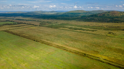 Fototapeta premium Aerial view of the country road and power lines near to the hill range, small coniferous forest and meadows agriculture at summer cloud sunny with horizon, yellow color