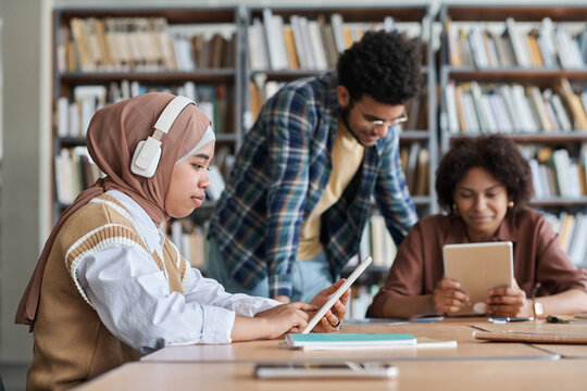 Muslim student in wireless headphones listening to online lesson on tablet pc while studying in the library with other students