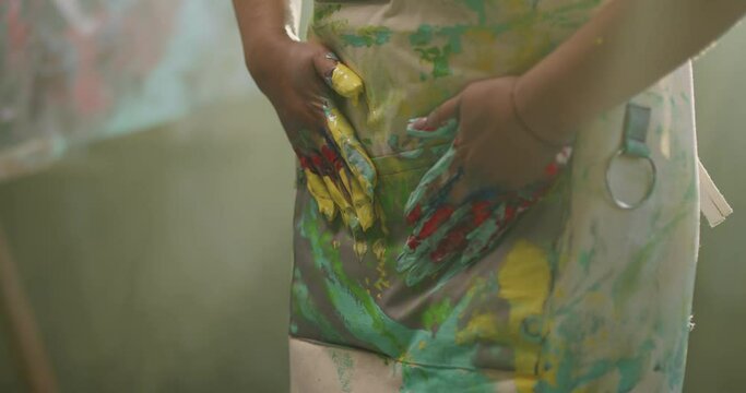 Artist millennial girl wipes her paint-stained hands on her apron in the rays of daylight. Modern artwork paint on canvas, creative, contemporary and successful fine art artist drawing masterpiece