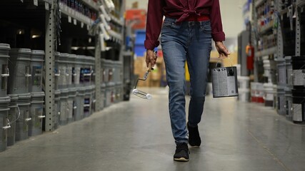 Woman shown from waist down, carrying can of paint and roller walking down paint aisle in hardware...