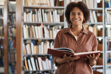 Portrait of African American student smiling at camera while reading book in library - obrazy, fototapety, plakaty