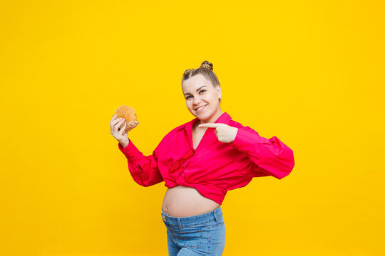 Funny Pregnant Young Woman Eating Burger. Pregnant Woman Holding Tasty Sandwich While Standing Isolated On Yellow Studio Wall. Junk Food Concept. Fast Food During Pregnancy.