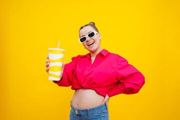 Cheerful pregnant woman in pink shirt holding tasty drink on isolated yellow background. A refreshing drink during pregnancy. A pregnant woman drinks water from a disposable glass