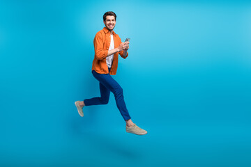 Fototapeta na wymiar Full length profile portrait of handsome excited guy use smart phone jumping rush empty space isolated on blue color background