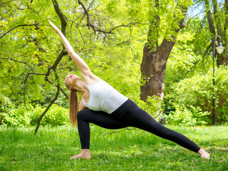 young pretty blond woman practicing yoga outdoor in the park in a sunny summer day, standing in parsvakonasana pose