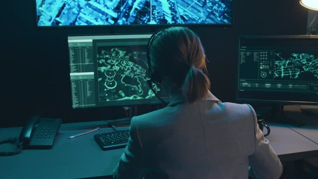 Back view of female Caucasian surveillance office worker sitting at desk in front of computer screens with monitoring program