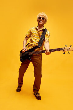 Full-length image of young smiling blonde man with curly hair in retro clothes playing guitar over yellow studio background. Concept of music, talent, hobby, entertainment, festival, performance, ad