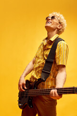 Happy, delightful, smiling man with blonde hair, in stylish clothes and sunglasses playing guitar on yellow studio background. Concept of music, talent, hobby, entertainment, festival, performance, ad