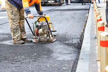 A road worker ramming a stretch of road around a sewer manhole with a gasoline vibrating plate.