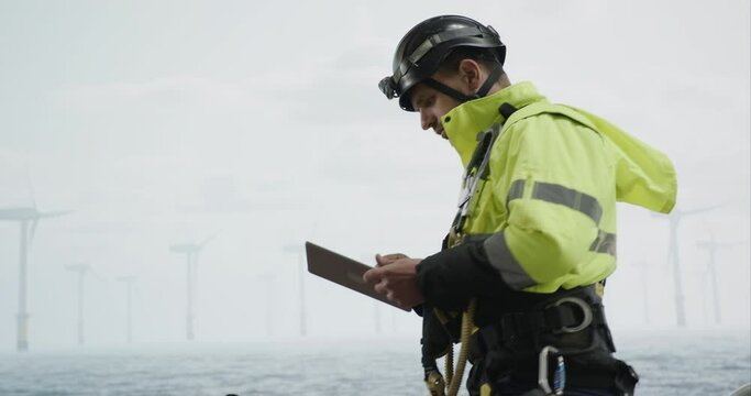 Maintenance engineer checking the data on tablet, approaching offshore wind turbine farm on a service vessel. Portrait of skilled professional preparing to work at high altitude