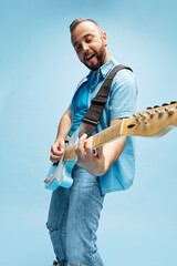 Stylish, positive, young bearded man in casual clothes playing electric guitar against blue studio...