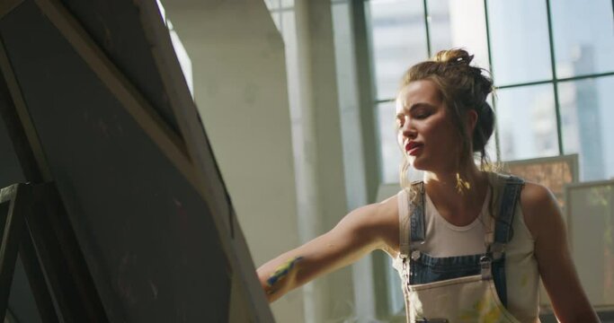 Millennial girl artist paints picture in loft studio using her hands to apply paint to canvas. Abstraction cinematic. Talented artist draws on the large canvas in light rays. Wipes hands on apron