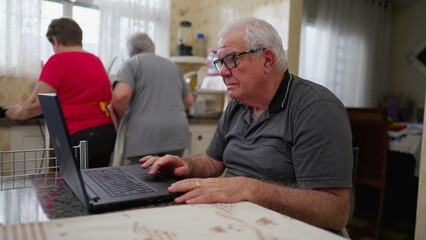 Older man in front of laptop using modern technology in domestic kitchen setting. Male caucasian senior engaged with computer at home