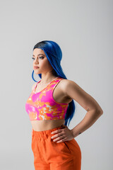 Obraz na płótnie Canvas fashion forward, tattooed young woman with blue hair posing with hands on hips in colorful clothes on grey background, individualism, modern style, urban fashion, vibrant color, female model