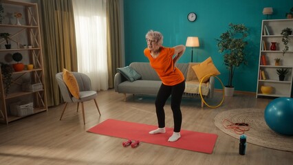 Chronic, agerelated, muscular back pain. An elderly woman, standing on a sports mat in front of dumbbells, holds her back. Retired woman holding lower back in pain while standing in living room.