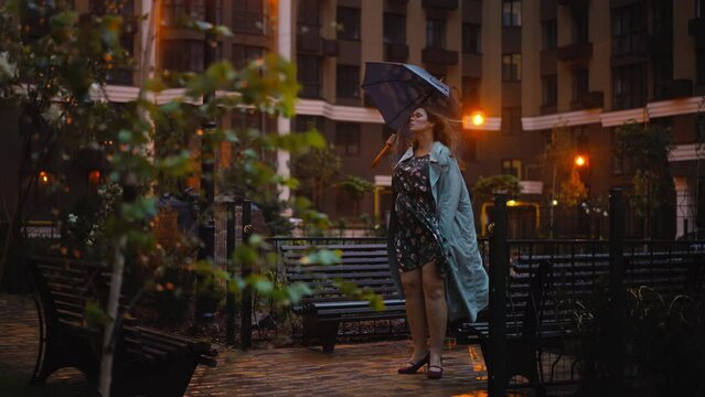 Wide shot thoughtful plus-size woman standing on windy rainy night in night city with umbrella. Beautiful Caucasian lady in dress coat and high-heels on illuminated street outdoors