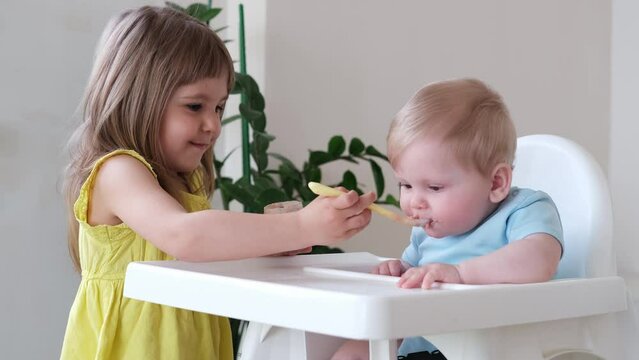 baby boy eating bland mashed food sitting on high chair, older sister feeding child 