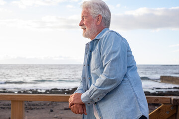 Portrait of beautiful senior bearded man in denim shirt standing outdoors at sea in sunset light looking away. Smiling old man relaxed enjoying retirement