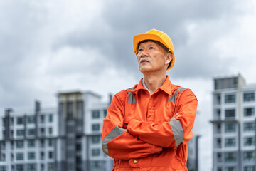 Serious and determined middle age Asian engineer in orange jumpsuit with hard hat crossing his arms while standing proudly in front of his multi-stories buildings