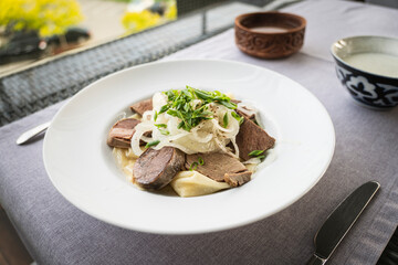Composition of tasty Kazakh national dish beshbarmak in a plate with kazy, flour tortillas, onion on a table in a restaurant