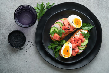 Healthy sandwich with bread or toast, smoked salmon, soft egg, cream cheese, cucumber, radish, black sesame and parsley on black plate. Delicious protein fish sandwich for breakfast. Smorrebrod.