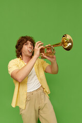 Portrait of young man with curly hair, talented musician playing trumpet against green studio...