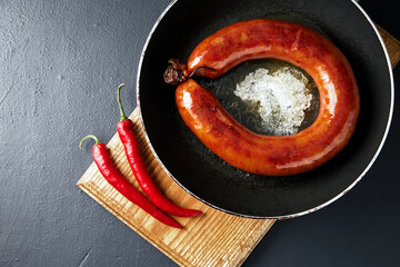 fried meat sausage in a cast iron pan and hot chili pepper on a cutting wooden board on a dark...