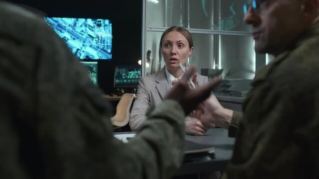 Over-the-shoulder shot of female civil officer arguing with two multiethnic co workers sitting at table in office at night during briefing in military hub