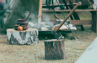 Axe and a cauldron on a stump in front of a campfire