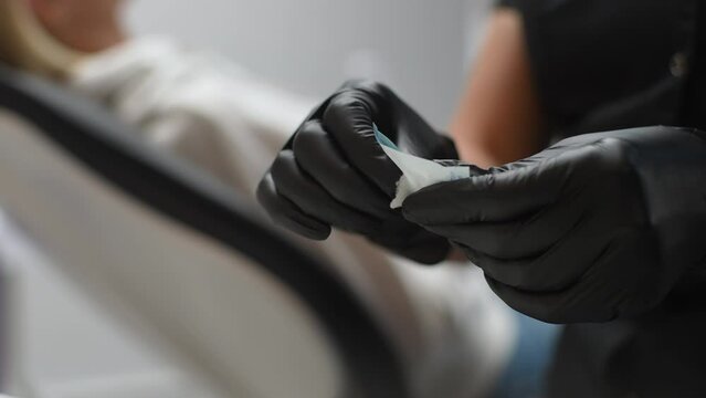 Close-up cropped shot of unrecognizable female dentist in rubber gloves opening package with new nozzle for polishing and grinding teeth of woman, sitting on dental chair on blurred background.