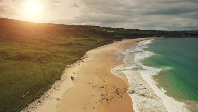 Aerial coast beach: sun, Atlantic Ocean, Northern Ireland, White Beach, Antrim county. People walking on sandy white shore with tranquil coastal wavy water. Cloudy summer scenery. Footage shot view