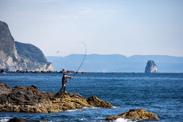 大海原を望む岩場で竿をキャスティングする釣り人