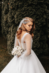 Photo of the bride in a wedding dress with sleeves, a beautiful hairstyle and a tiara on her head, holding a bouquet of flowers behind her back. © Vasil