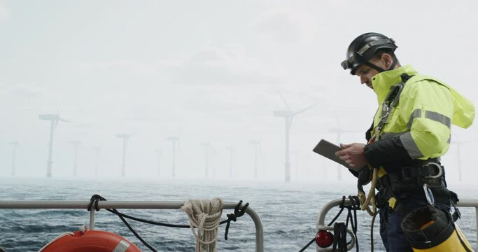Maintenance engineer checking the data on tablet, approaching offshore wind turbine farm on a service vessel. Portrait of skilled professional preparing to work at high altitude