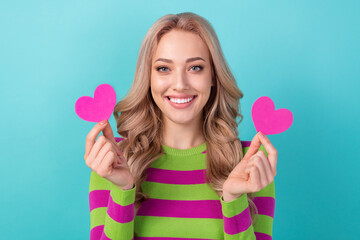 Photo portrait of attractive young woman hold two little pink hearts cards wear trendy striped clothes isolated on cyan color background