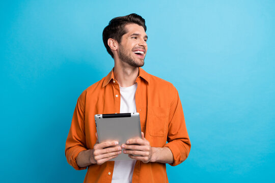Photo of positive excited guy wear orange shirt texting modern device looking empty space isolated blue color background